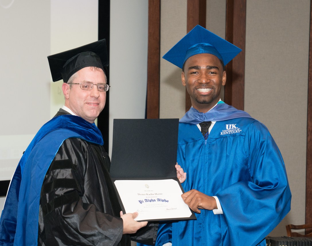 Dexter Horne wearing a blue graduation robe and smiling as he receives an academic award from the Martin School's program director.